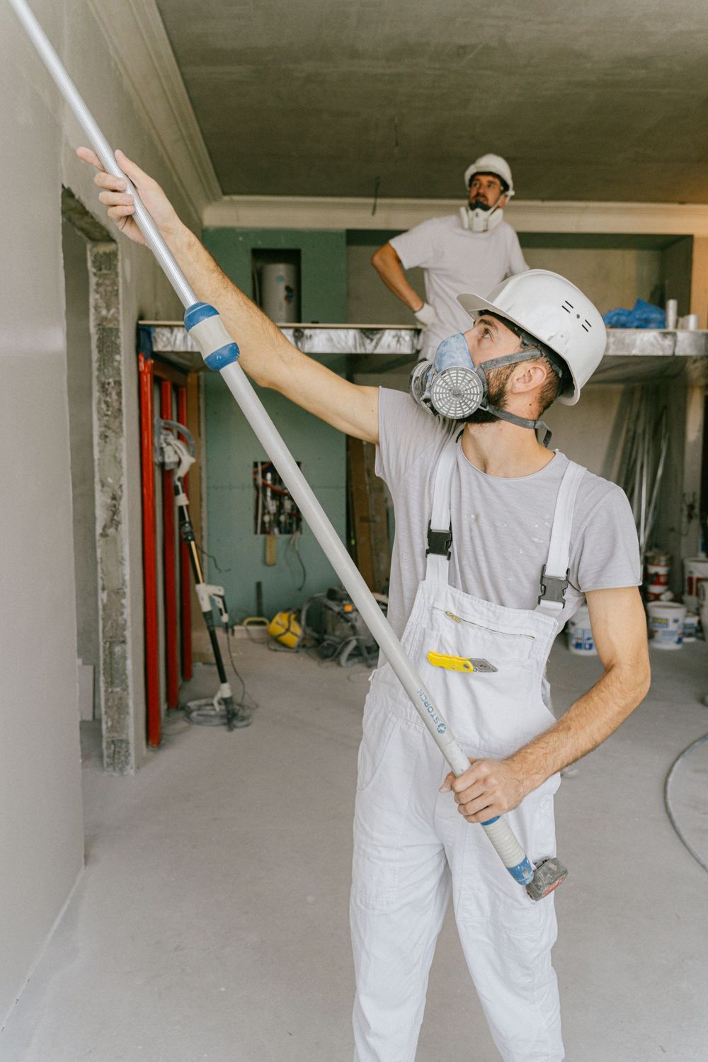 Vakman in witte overall werkt met een lange stang aan een plafond in een kale ruimte